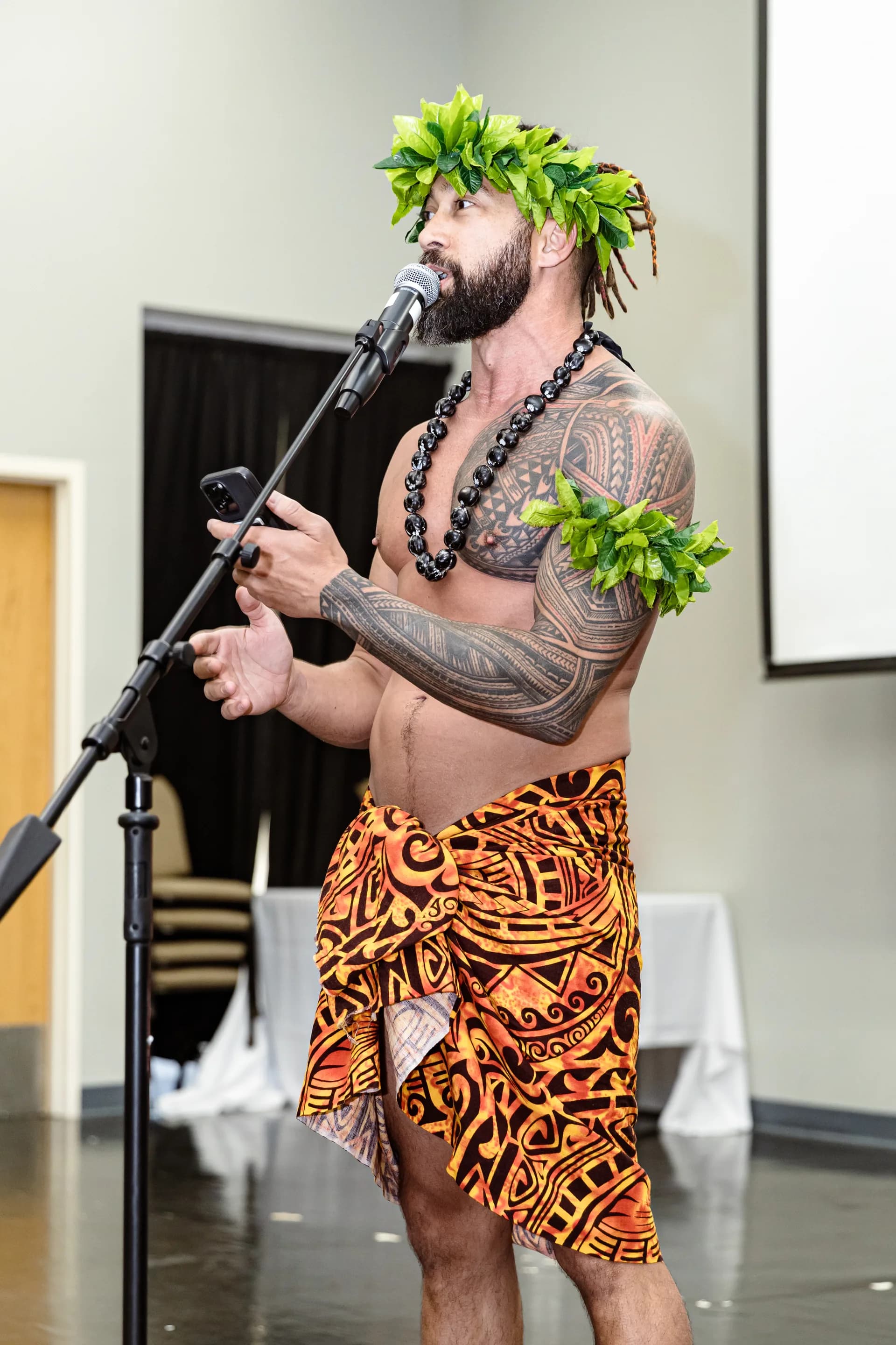 A tattooed man in an orange and black sarong sings into a microphone on a stand, a green leaf headpiece and armband marking him as a Polynesian performer.