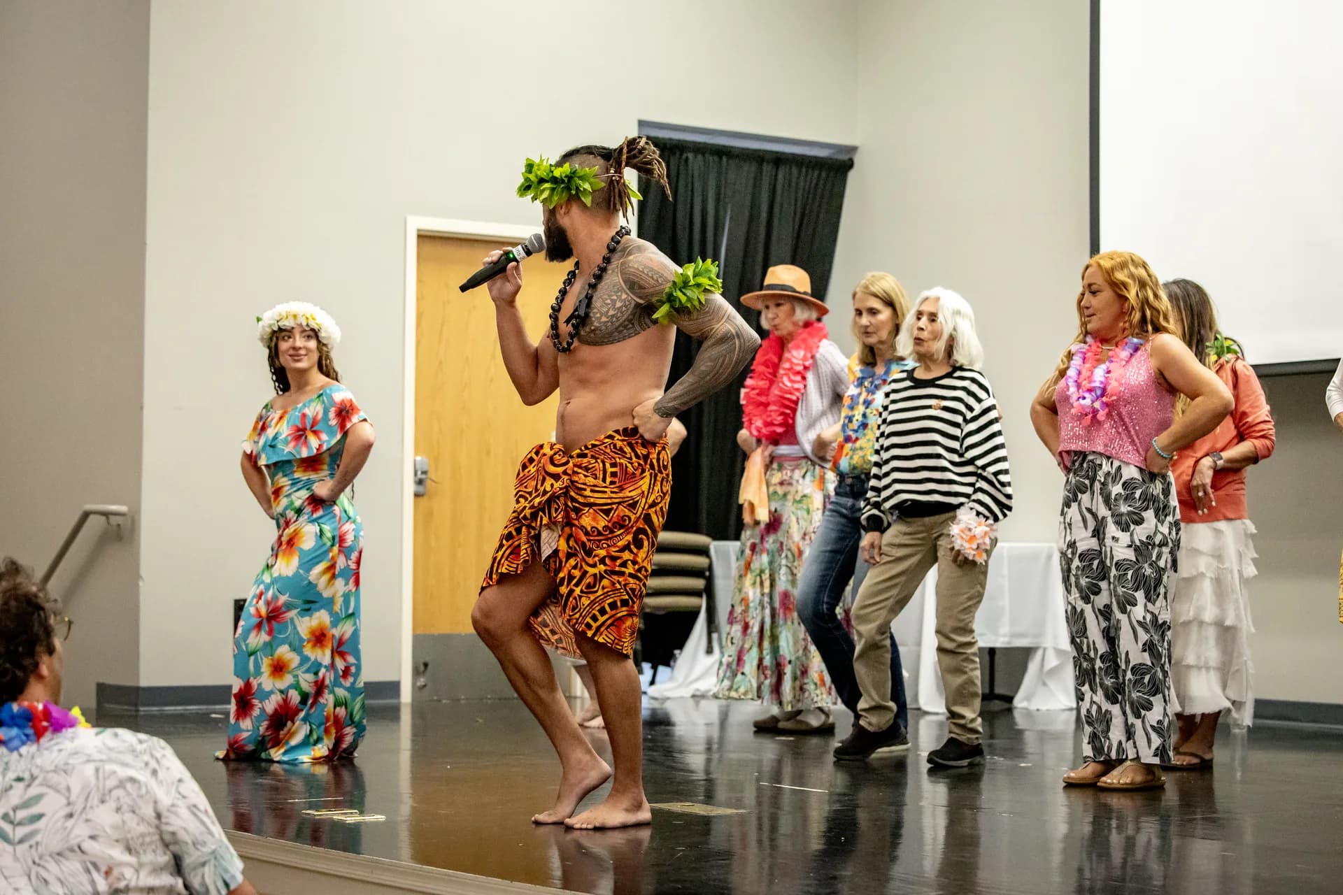 The same performer mid-step on a stage holding a handheld microphone, guests wearing leis watching from behind him.