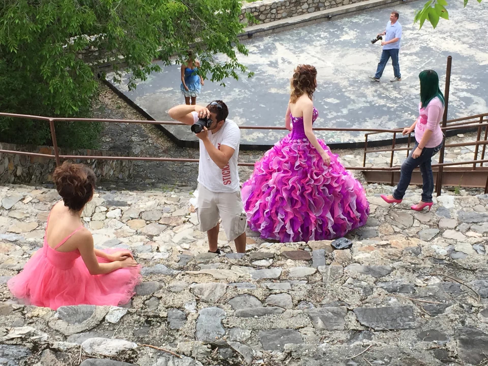 A younger Joseph in a white t-shirt and cargo shorts raises a professional camera to his eye on stone stairs, photographing a woman in a bright pink ballgown while other models and a second photographer work the scene behind him.