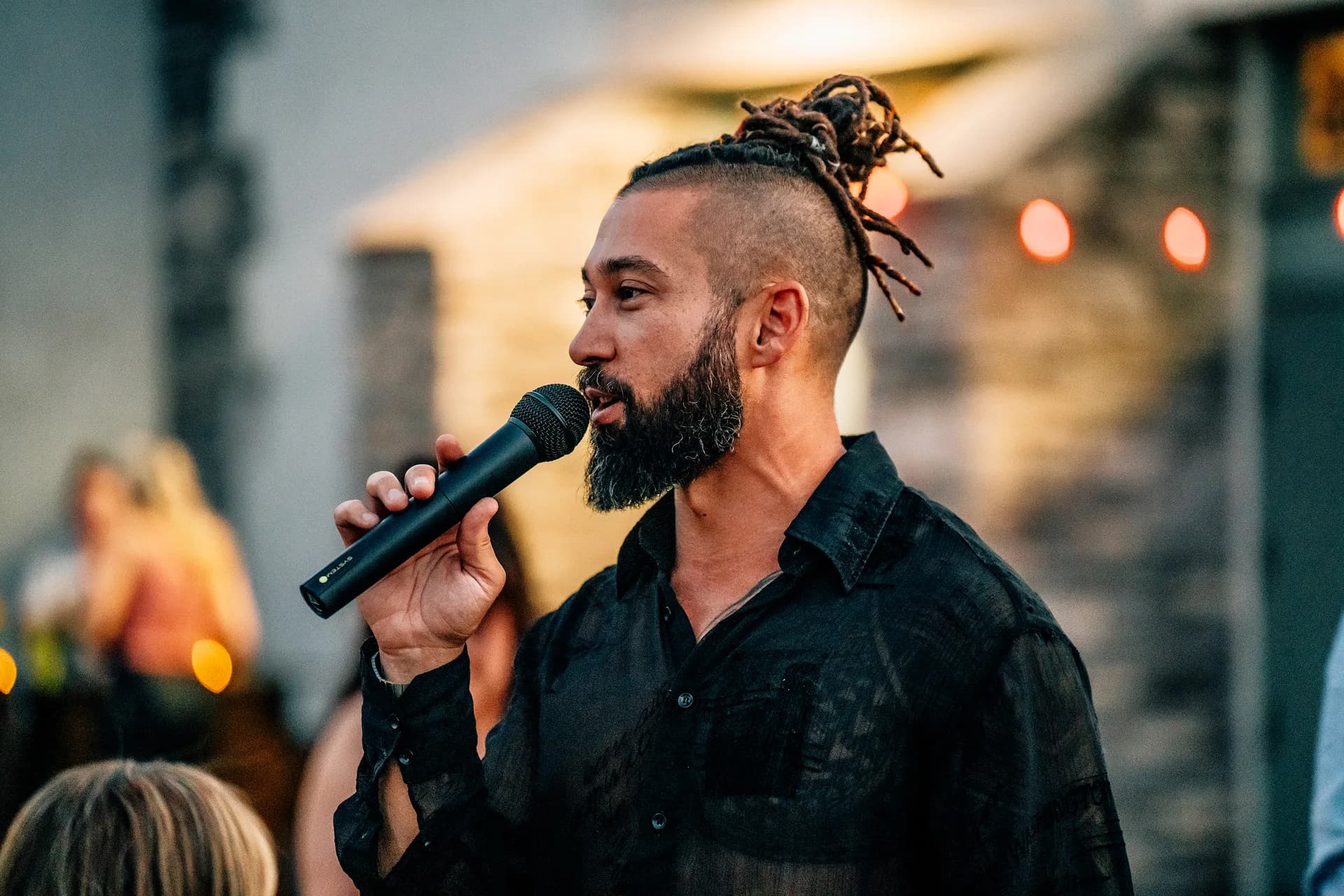 Joseph in a black shirt speaks into a handheld microphone at an outdoor evening event.