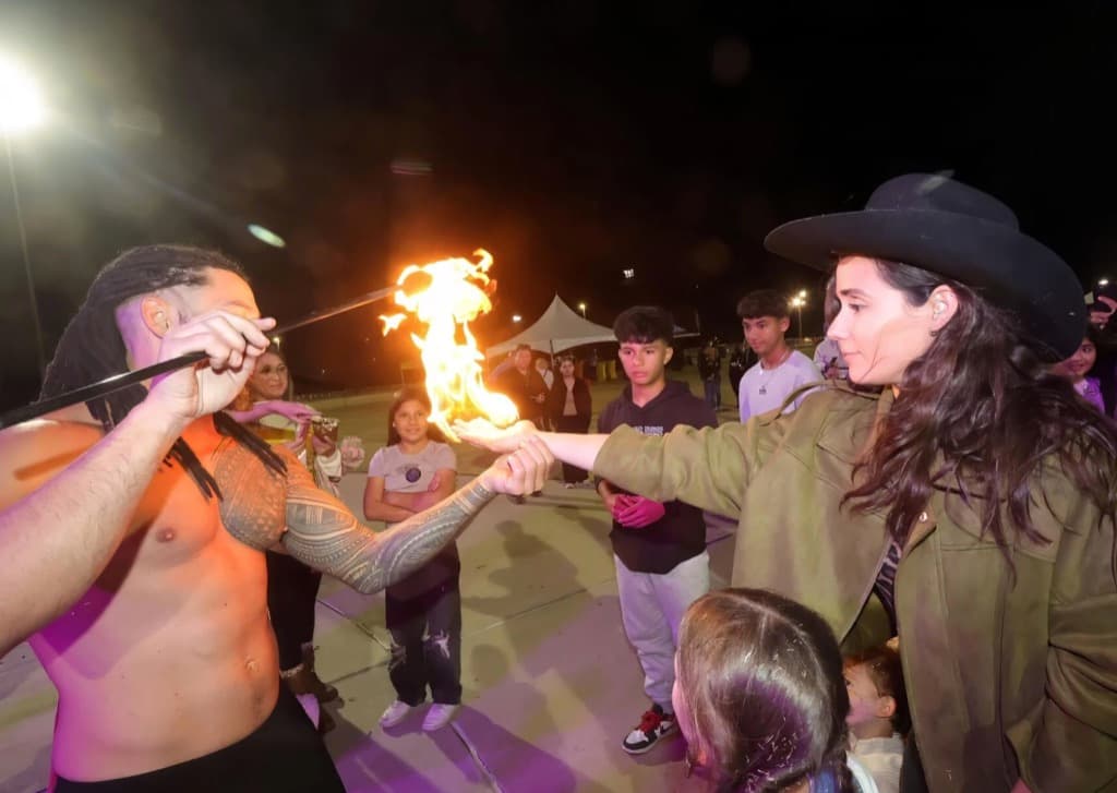 A fire performer holds a flaming staff at night while a woman in a hat touches the flame and a crowd watches.
