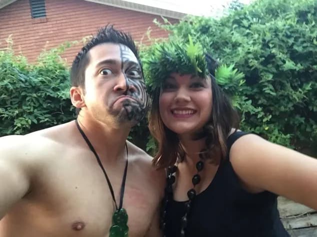 A man with tribal face paint and a woman wearing a green leaf crown pose together outdoors.