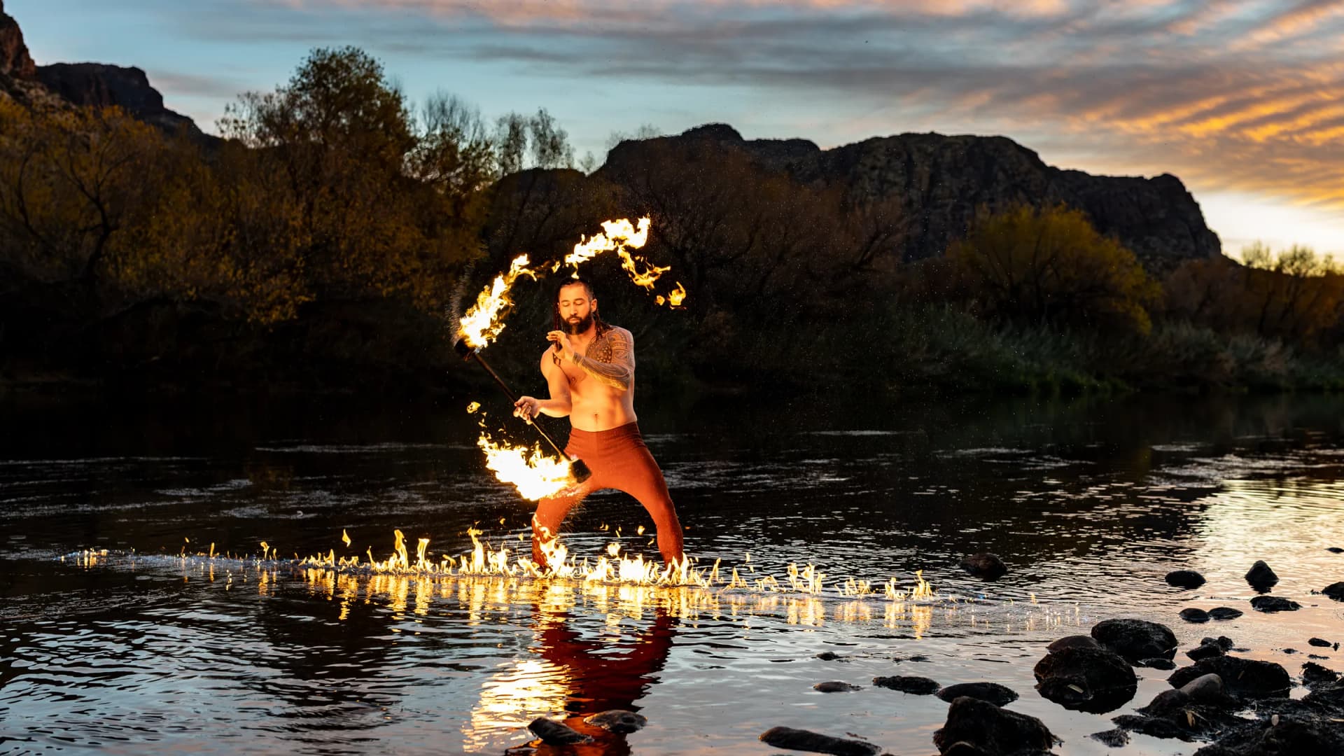 A fire performer swings a flaming torch while standing in a shallow river at sunset, mountains beyond.