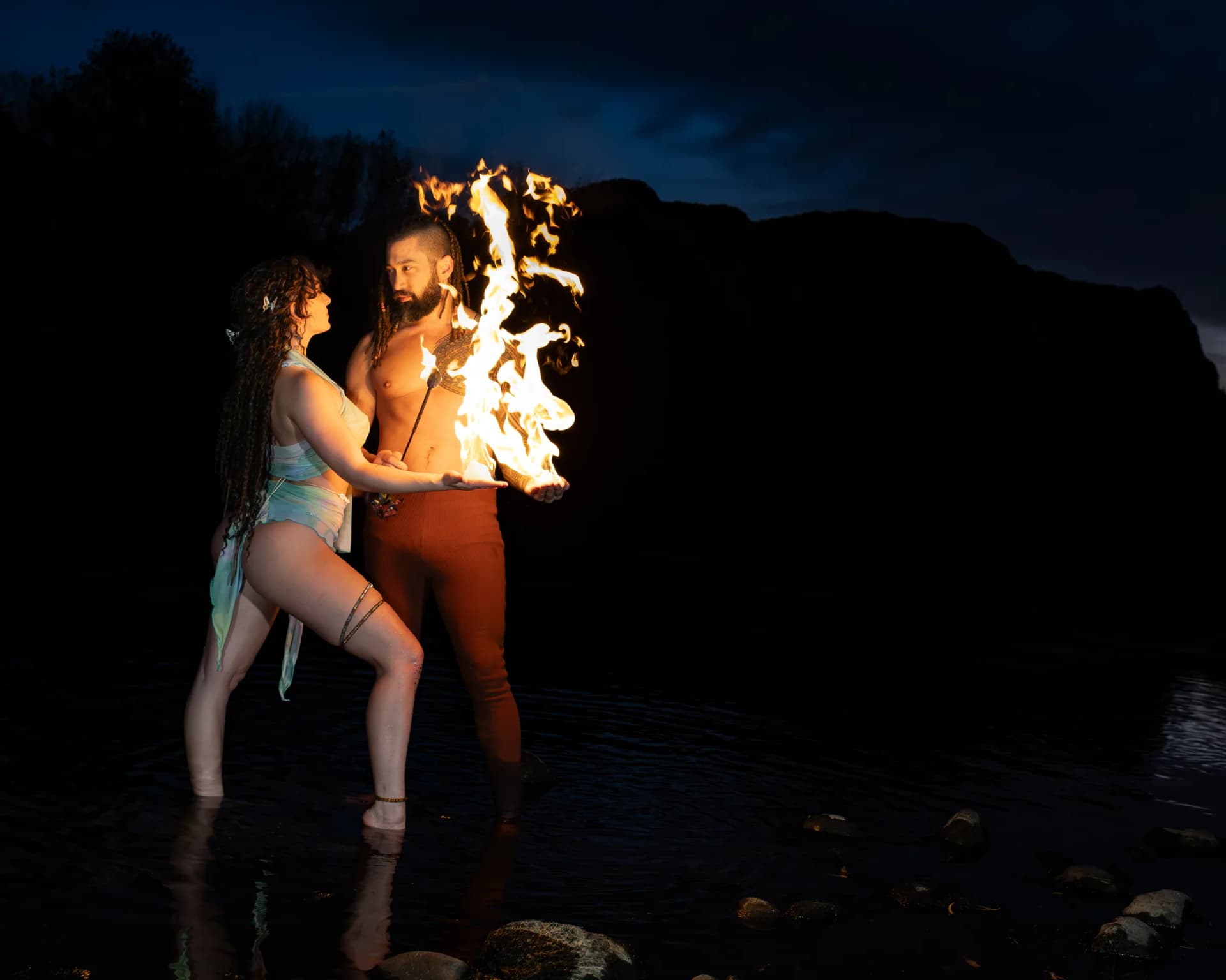 A man and woman stand close at night as flames rise from his cupped hands between them.