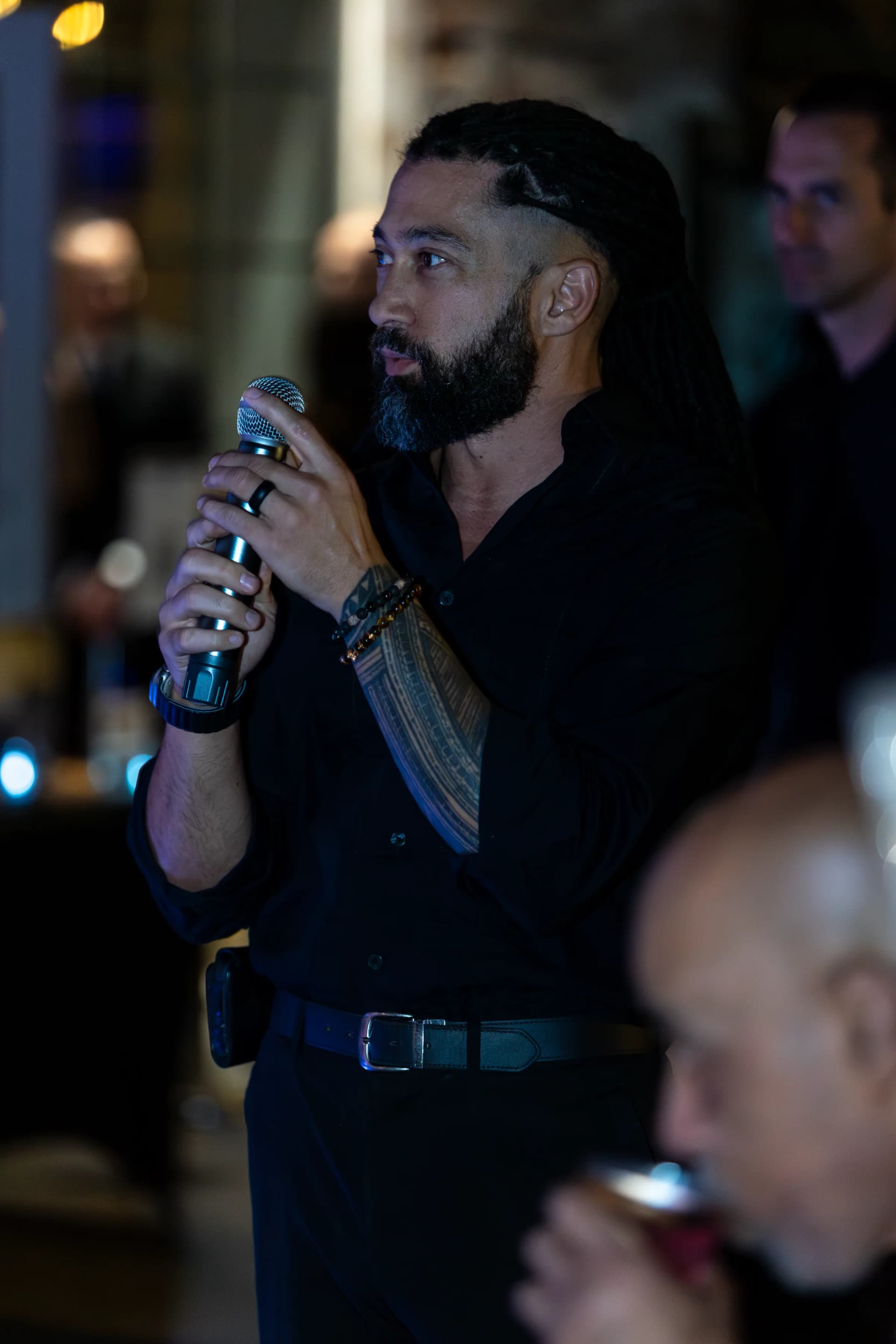 A bearded man with a tattooed arm speaks into a microphone at an indoor gathering.