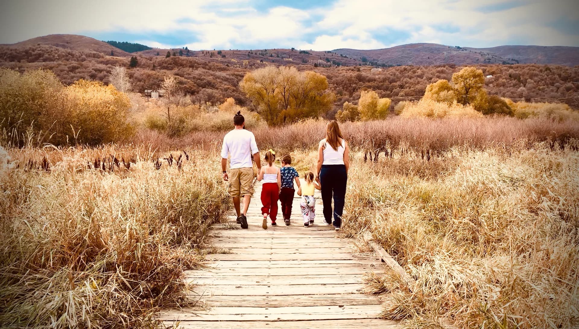 A father and mother walk hand in hand with three children along a wooden boardwalk through golden grass.